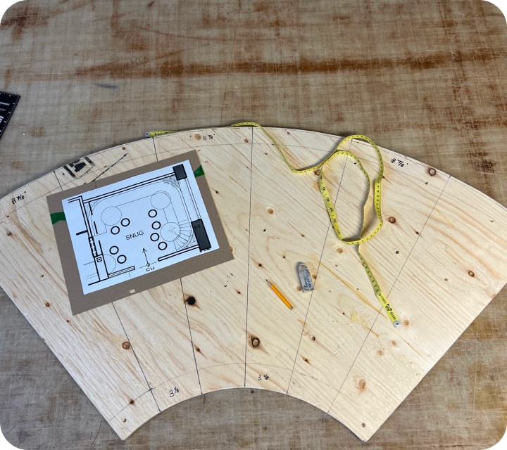 Top view of a quarter-circle plywood panel with lines drawn to separate the top into six identical sections. A flexible measuring tape, an eraser, a pencil, and a sketch of the layout plan for the future bench seat are placed on it.