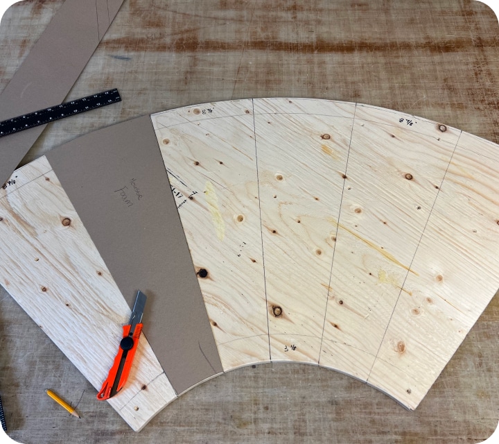 Top view of a quarter-circle plywood panel with lines drawn to separate the top into six identical sections. A piece of cardboard measuring exactly one of the six sections, a pencil, and an exacto ruler are placed on top.