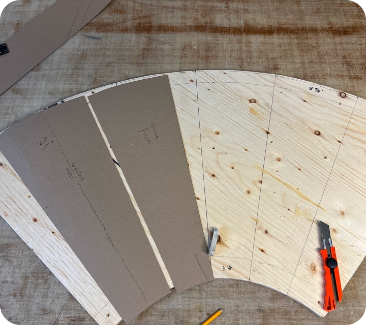 Top view of a quarter-circle plywood panel with lines drawn to separate the top into six identical sections. A cardboard box with the exact dimensions of one of the six sections, an attached cardboard box indicating a seam line for a ripper, a pencil, an eraser, and an exacto knife are placed on top.