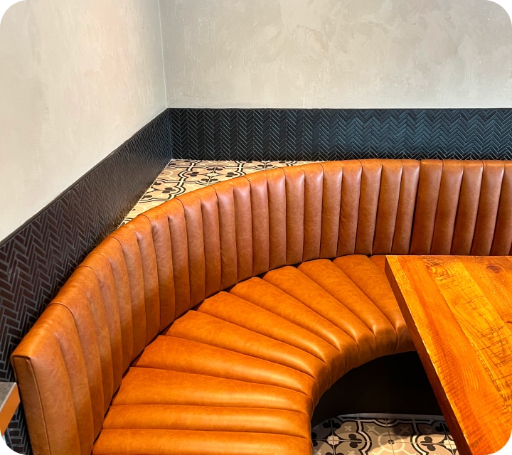 Partial overhead view of a rounded corner of a restaurant dining room banquette. The seat and back are made of padded and joined flutes (tubes). Rounded corner. Caramel-colored faux leather. Banquette custom-made according to the customers' choices and preferences. Réno-Montréal collaboration