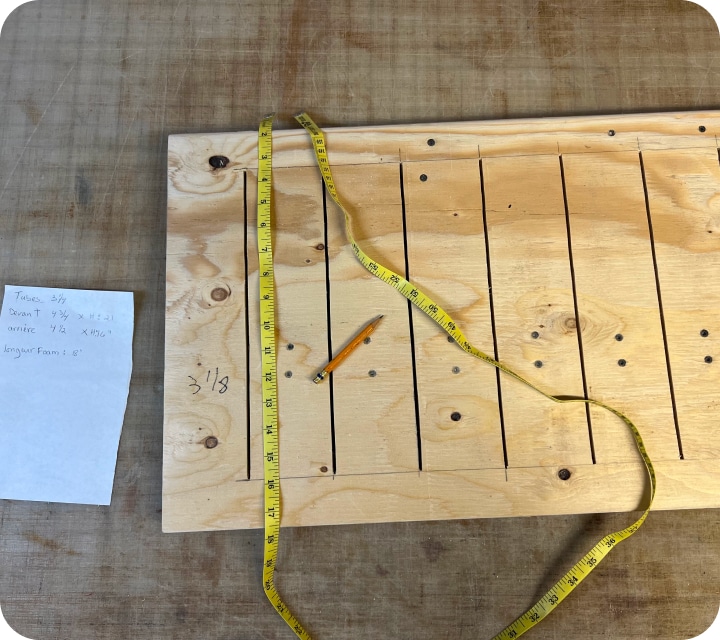 Close-up top view of a piece of wood with slots (openings), a tape measure lying on the floor, and a pencil. Some illegible notes on a piece of paper to the right.