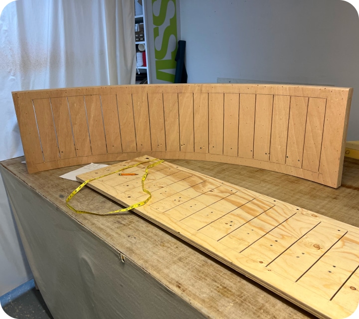 A view of a rounded plywood panel raised on a table. In front of it is a plywood panel with slots, also lying on the table. A measuring tape and a pencil are placed on it.