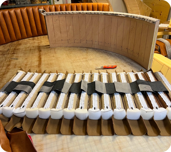 Rear view of a set of padded tubes that will cover a rounded backrest. Each tube is joined to its neighbors to form an assembly. Fabric tabs are sewn to pass through the slots in the panel to be upholstered and help hold the seam assembly firmly against the rounded shape of the backrest.
