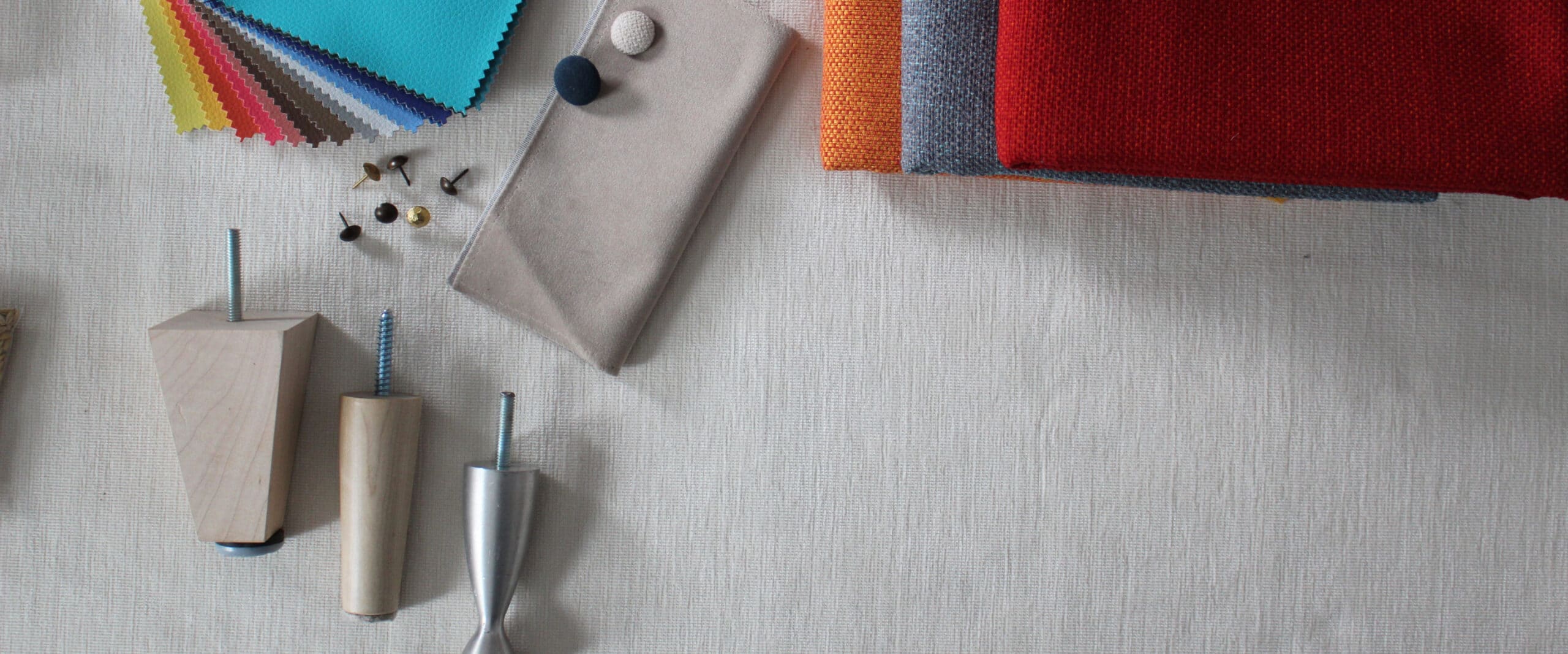 Top view of an arrangement of materials and coverings for upholstered furniture: at the bottom left, a set of two legs in wood and brushed aluminum metal, above some scattered decorative nails and some examples of coverings in various colors presented in a fan and in a pile at the top right.